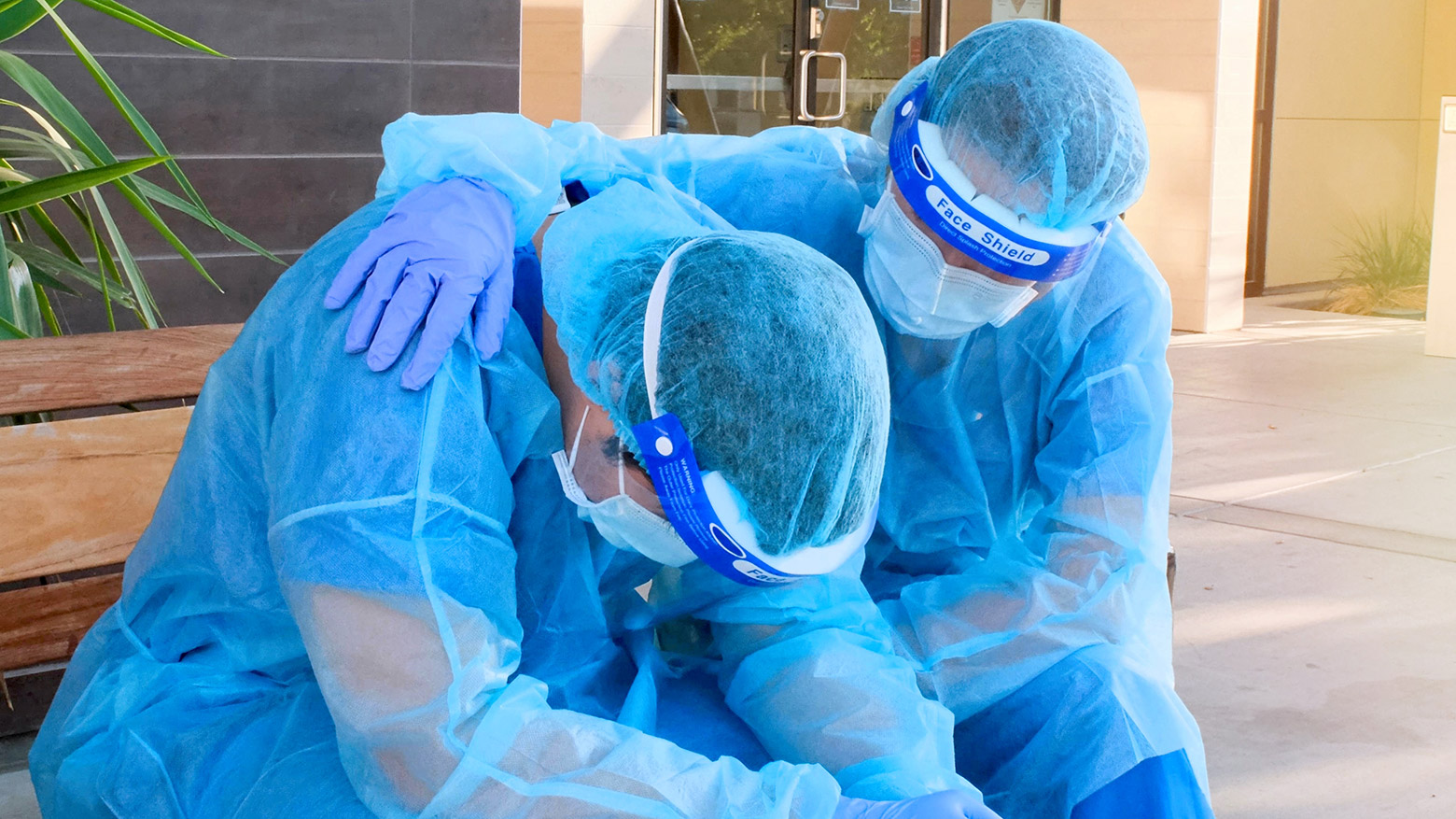 Two doctors in PPE on a bench. One is slumped over with his head down, while the other comforts him.